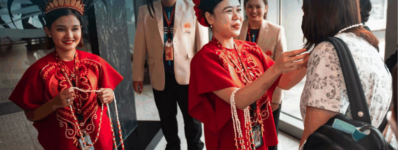 Airport staff in red traditional dress and feathered headdresses welcoming arriving passengers with shell necklaces