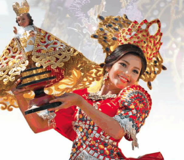 Smiling Sinulog dancer in a red sequined costume and gold headdress holding a Santo Niño image